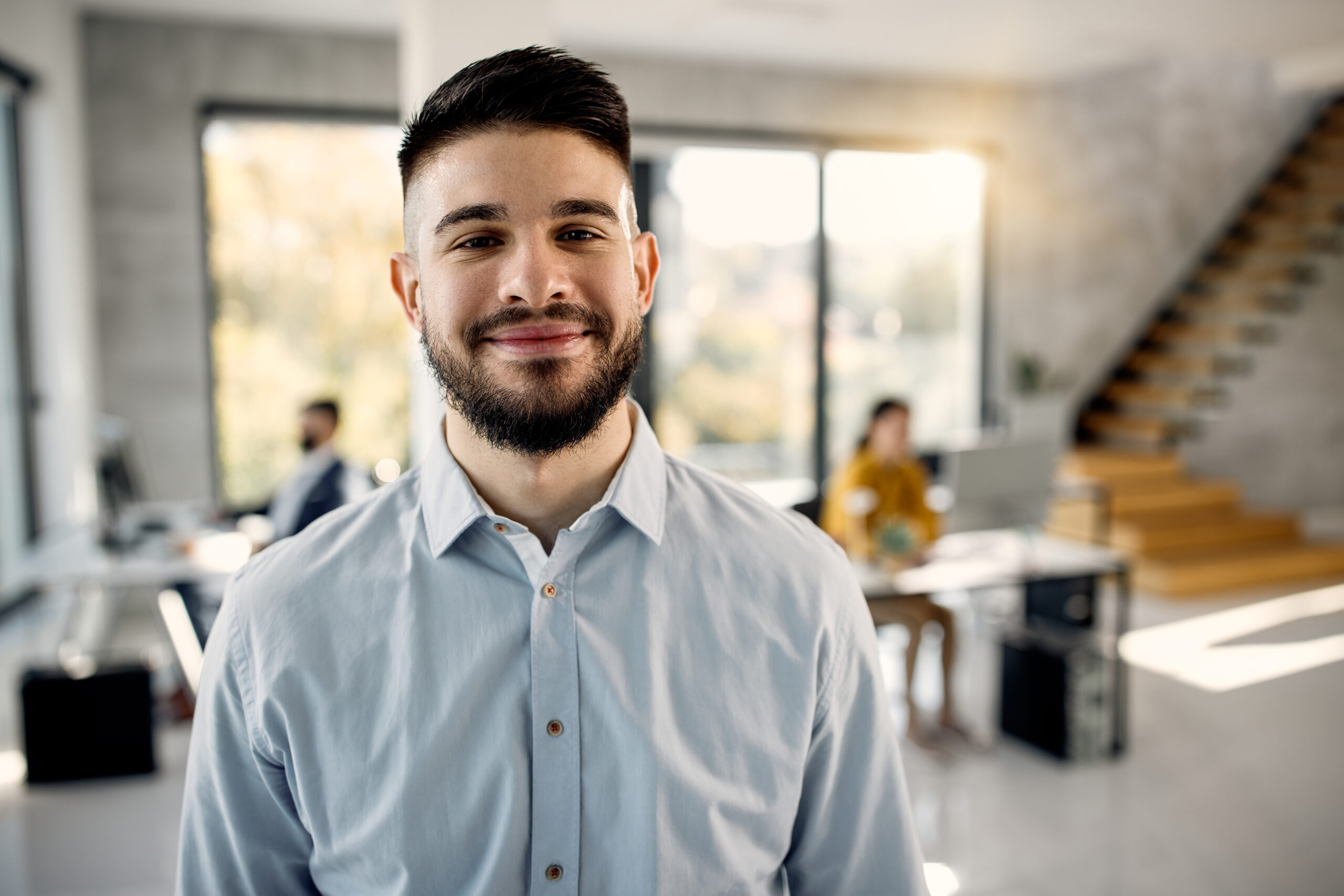 Young happy entrepreneur standing in the office and looking at camera. Hid colleagues are working in the background.